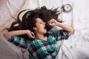 "Woman sleeping peacefully in bed with sunlight peeking through window blinds. The alternative text describes the photo that accompanies an article about optimizing your sleep schedule."