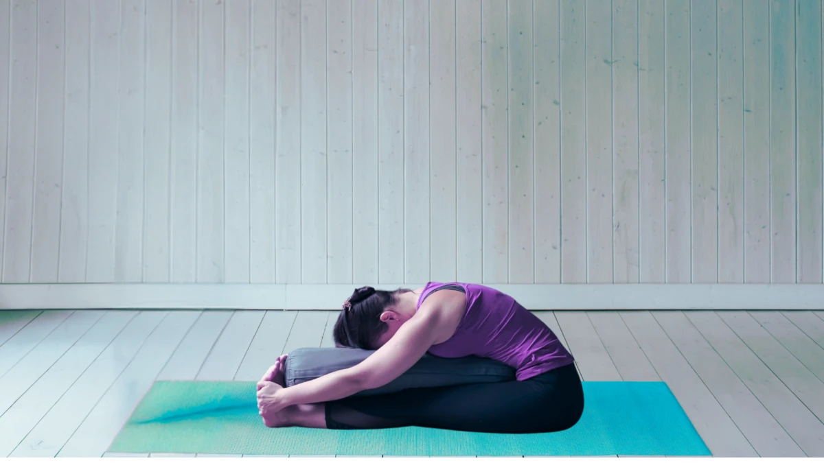 Person sitting with legs extended, leaning over a vertically positioned yoga bolster placed between the thighs